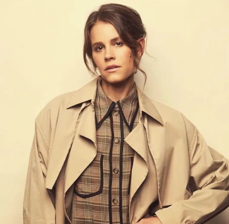 Portrait of a woman wearing a plaid shirt and beige trench coat against a solid neutral backdrop