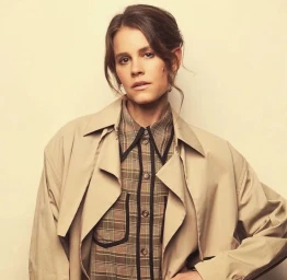 Portrait of a woman wearing a plaid shirt and beige trench coat against a solid neutral backdrop