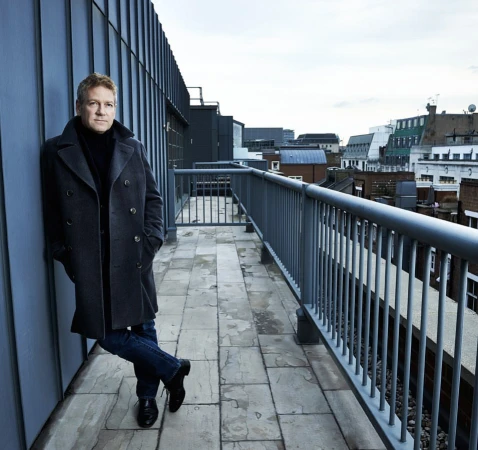 Portrait of Kenneth Branagh leaning against a dark wall on a rooftop balcony wearing a grey double breasted coat