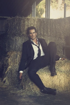 Man in a dark suit and loose tie leaning against large bales of hay in a rustic barn setting by Brian Bowen Smith