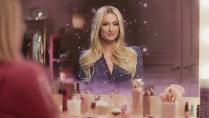 Close-up of a woman with long blonde hair in a purple shirt, reflecting in a vanity mirror surrounded by perfume bottles.