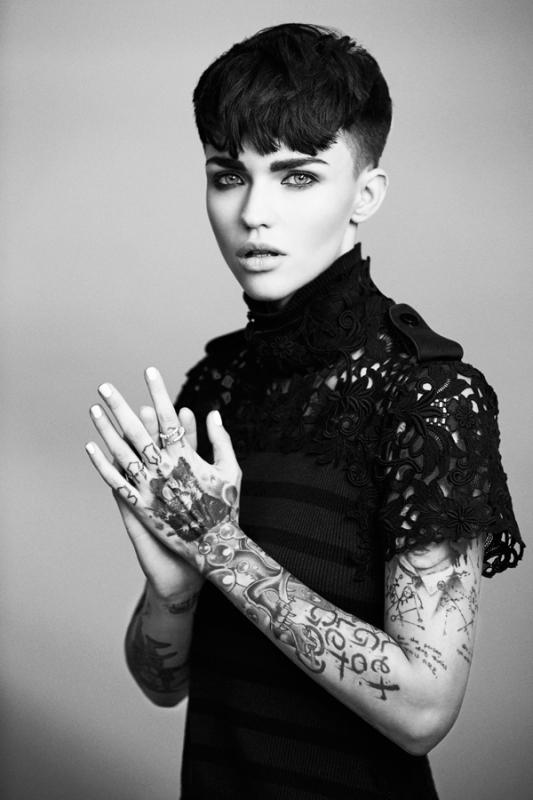 Black and white portrait of a young woman with dark hair, tattoos on her arms and hands, and wearing a high-necked, lacy top.