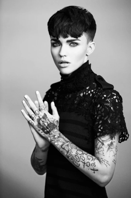 Black and white portrait of a young woman with dark hair, tattoos on her arms and hands, and wearing a high-necked, lacy top.