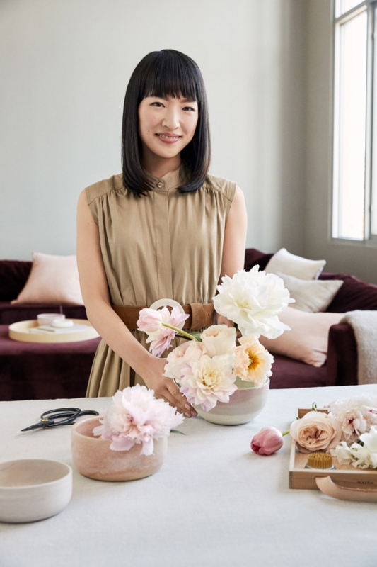 Adult woman arranges pale pink and white peonies in decorative vases on a white tablecloth with a neutral-toned sofa and pillows in the background.
