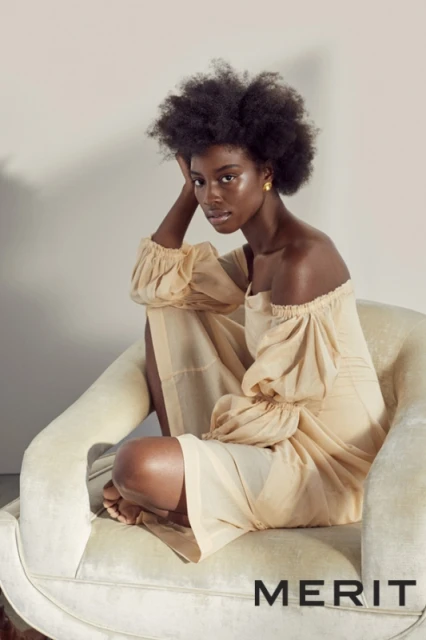 Elegant dark-skinned woman poses in off-the-shoulder dress on an armchair against a neutral backdrop, showcasing fashion and beauty.