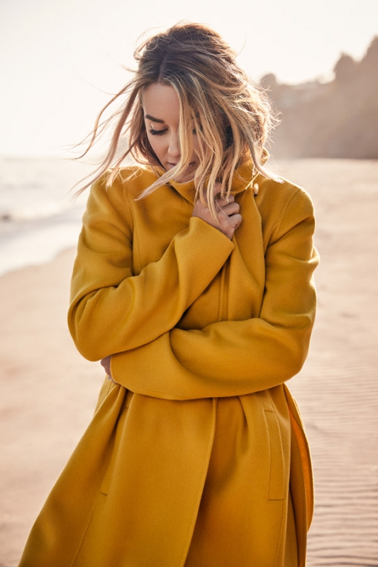A woman in a yellow coat stands on a sandy beach with arms crossed, looking down, with soft, warm lighting and a blurred background.