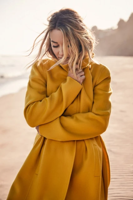 A woman in a yellow coat stands on a sandy beach with arms crossed, looking down, with soft, warm lighting and a blurred background.
