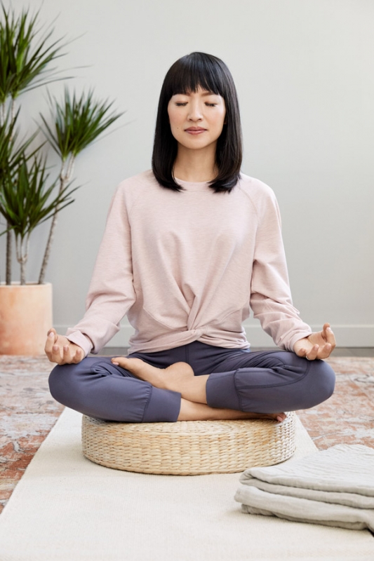 Woman meditates indoors in a lotus position on a woven cushion. She wears casual athletic clothing with plants and a light wall backdrop.