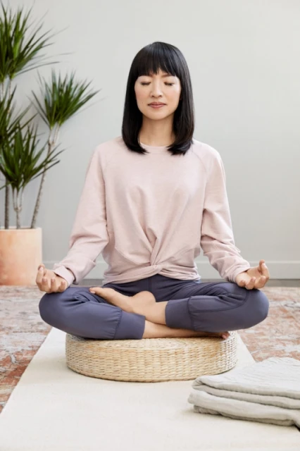 Woman meditates indoors in a lotus position on a woven cushion. She wears casual athletic clothing with plants and a light wall backdrop.