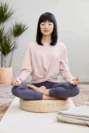 Woman meditates indoors in a lotus position on a woven cushion. She wears casual athletic clothing with plants and a light wall backdrop.