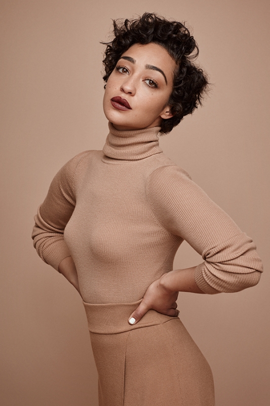 Elegant studio portrait of a woman with curly short hair wearing a neutral turtleneck and pants, posing with hands on her waist against a neutral backdrop.