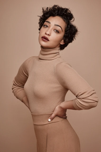 Elegant studio portrait of a woman with curly short hair wearing a neutral turtleneck and pants, posing with hands on her waist against a neutral backdrop.