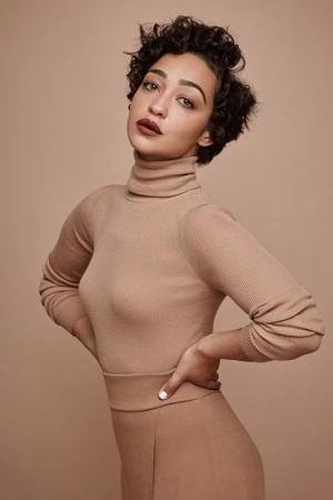 Elegant studio portrait of a woman with curly short hair wearing a neutral turtleneck and pants, posing with hands on her waist against a neutral backdrop.
