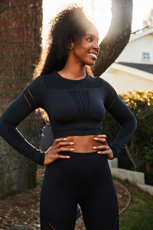 Smiling Black woman in black crop-top workout gear poses in a sunny outdoor setting, showing off her athletic build and curly hairstyle.