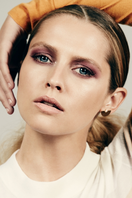 Close-up portrait of a woman with purple eyeshadow and a white top against a neutral background by Justin Coit.