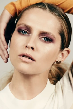 Close-up portrait of a woman with purple eyeshadow and a white top against a neutral background by Justin Coit.