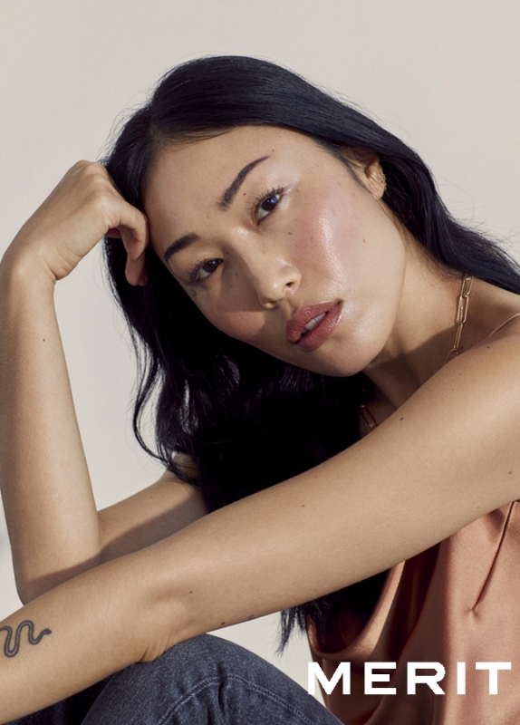 Portrait of an Asian woman with dark hair, wearing a tank top, posing indoors with soft lighting in a beauty advertisement for Merit.