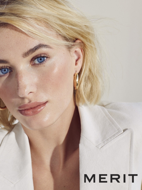 Close-up of a blonde woman with blue eyes wearing gold earrings and a white blazer against a light background; beauty and fashion.