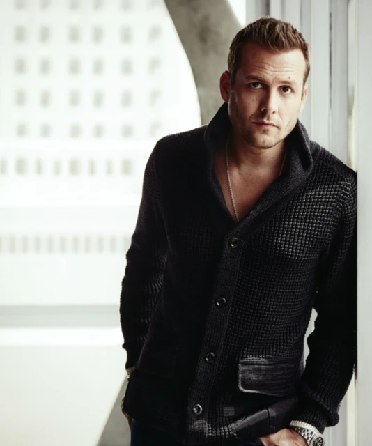 Portrait of a stylish adult man in a dark cardigan leaning against a wall with a neutral expression, set against an office backdrop.
