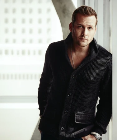 Portrait of a stylish adult man in a dark cardigan leaning against a wall with a neutral expression, set against an office backdrop.