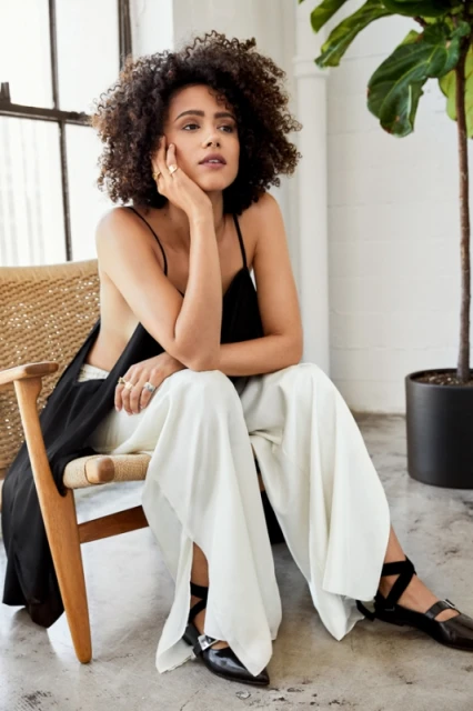 Woman with curly hair in spaghetti strap top and palazzo pants sitting in a chair indoors with natural lighting from a window.
