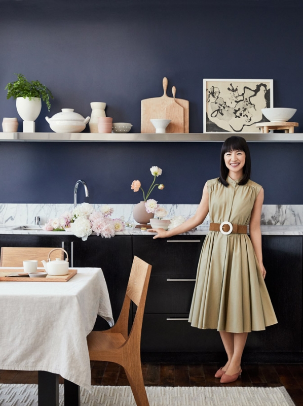 Marie Kondo smiles in a minimalist kitchen with dark blue walls, a light wood table setting, and assorted kitchenware on display.