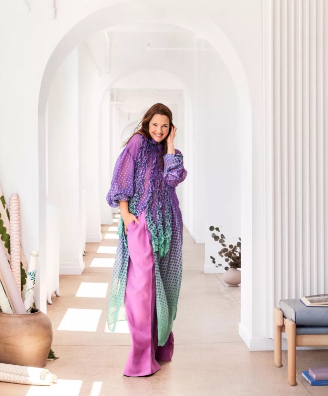 A cheerful woman poses in a light-filled hallway wearing a stylish, flowing purple and pink dress, exuding a relaxed and confident vibe.