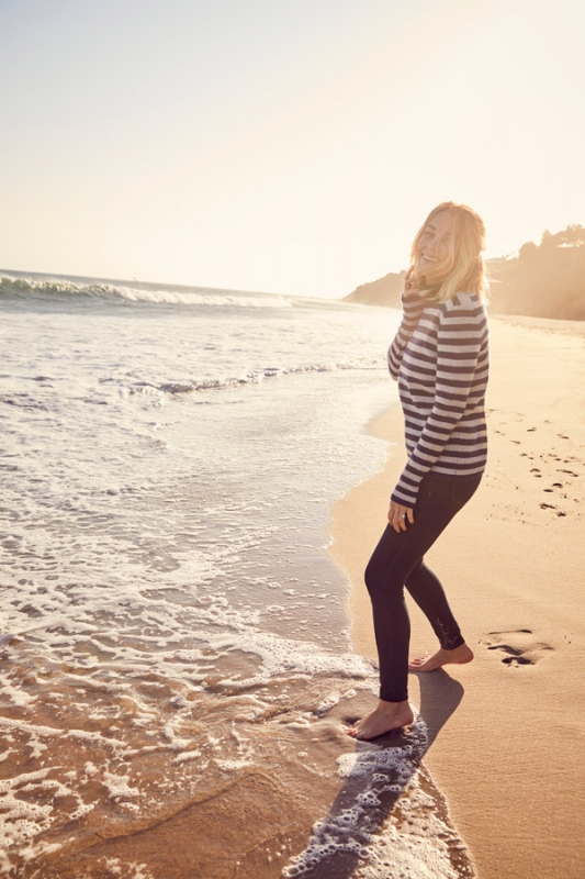 Woman in striped sweater standing barefoot on a sandy beach near ocean waves at sunset