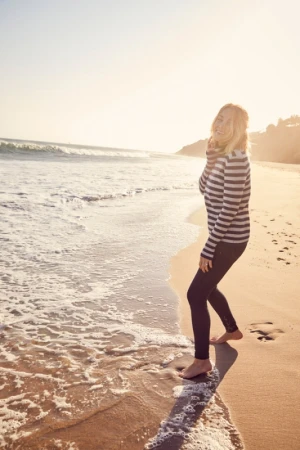 Woman in striped sweater standing barefoot on a sandy beach near ocean waves at sunset