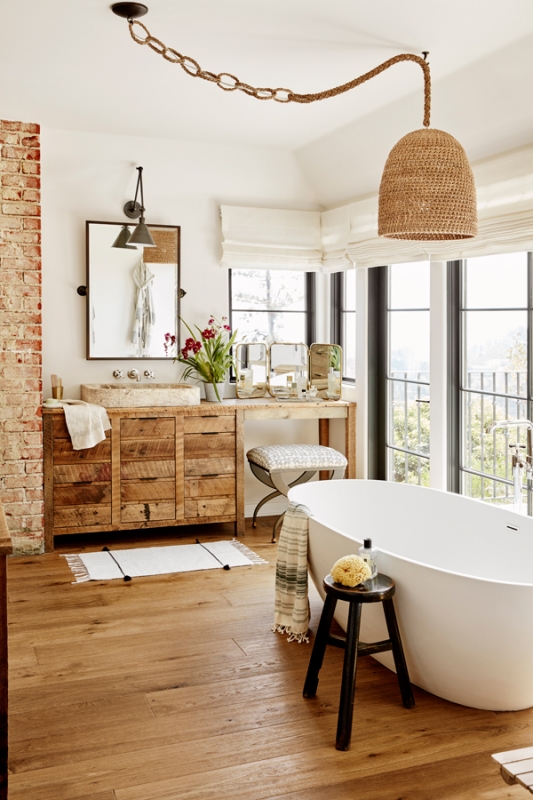 Bright and airy bathroom interior featuring a freestanding bathtub, rustic vanity, woven pendant light, and natural wood flooring.