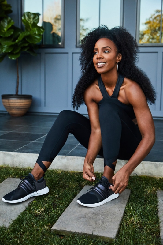 Athletic woman with dark curly hair tying her black and gray sneaker on a stone pathway with a gray building in the background.