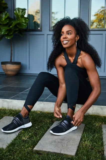 Athletic woman with dark curly hair tying her black and gray sneaker on a stone pathway with a gray building in the background.