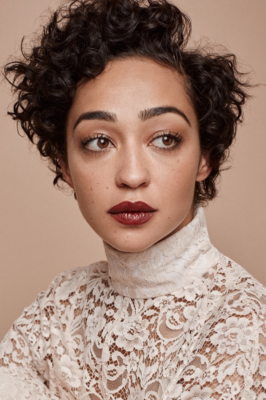 Close-up portrait of a woman with short dark curly hair wearing a high-neck white lace top and dark lipstick against a beige background