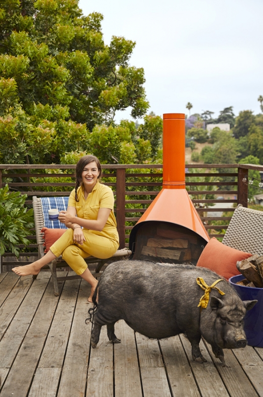Woman sitting on a wooden chair on a deck with a pig, an orange outdoor fireplace, trees, and a neighborhood in the background.