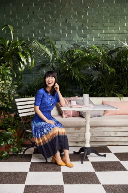 Woman in blue dress seated at table on checkered floor, surrounded by indoor plants, creating a vibrant and stylish indoor portrait.