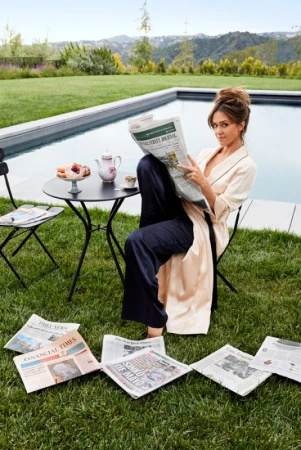 Jessica Alba reading a newspaper by an outdoor swimming pool in a white robe and dark pants, photographed by Justin Coit.