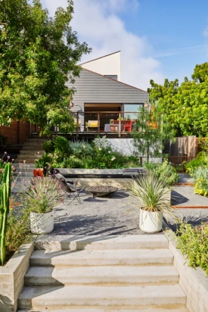 Exterior view of a modern house with a deck, landscaped garden, and staircase leading to a gravel courtyard with a fire pit and chair.