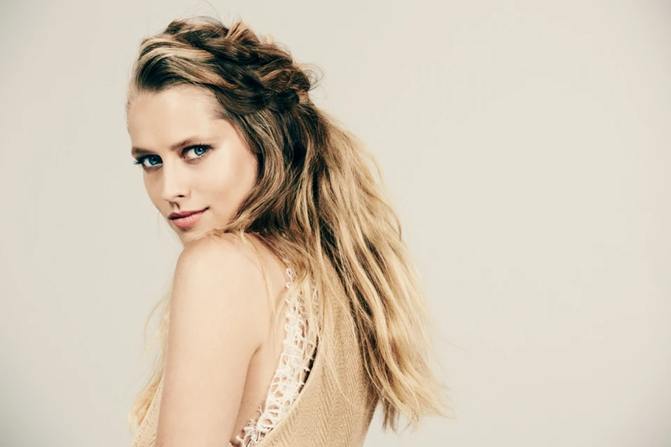 Close-up studio shot of a woman with blonde hair and blue eyes looking over her shoulder, wearing a tan top with lace details.