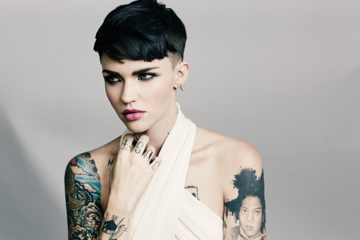 Close-up portrait of Ruby Rose with short black hair and tattoos, wearing a white wrap top against a neutral gray backdrop.
