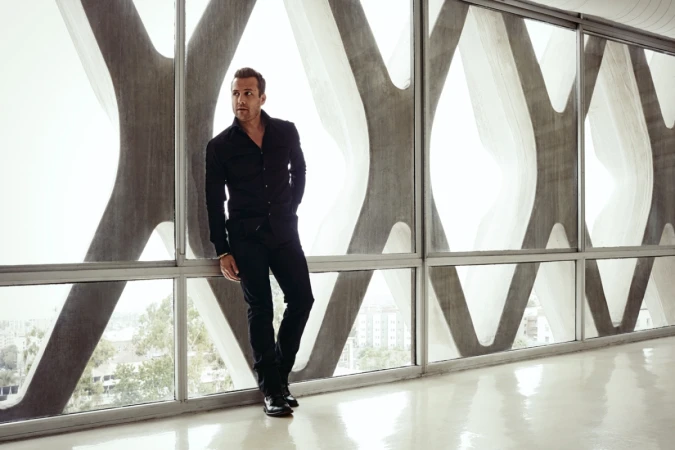 An adult male poses in all-black attire against a geometric window. He is seen in full shot with natural lighting indoors.
