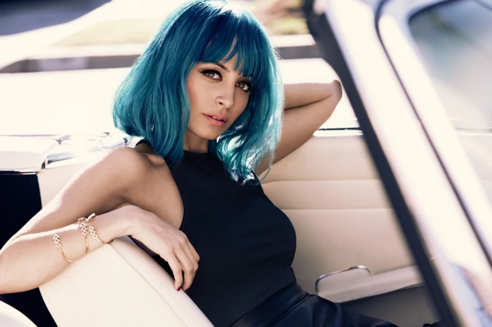 Stylish woman with blue hair and gold bracelets poses inside of a car. She wears a black top and skirt with natural light.