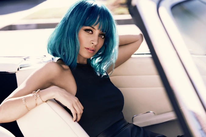 Stylish woman with blue hair and gold bracelets poses inside of a car. She wears a black top and skirt with natural light.