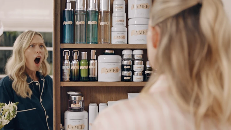 Blond woman in pajamas with surprised expression reflected in mirror, La Mer skincare products displayed on wooden shelf in background.