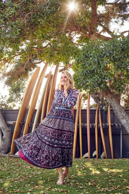 Barefoot woman twirls in a bohemian dress among surfboards in a sunlit outdoor setting with trees and a wooden fence.