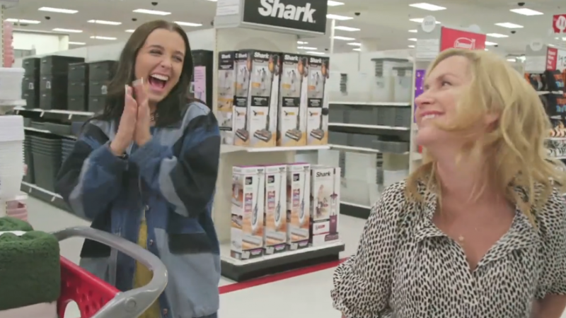 Two adult females stand inside a retail store, one clapping and smiling with a shopping cart, the other looking towards the first.
