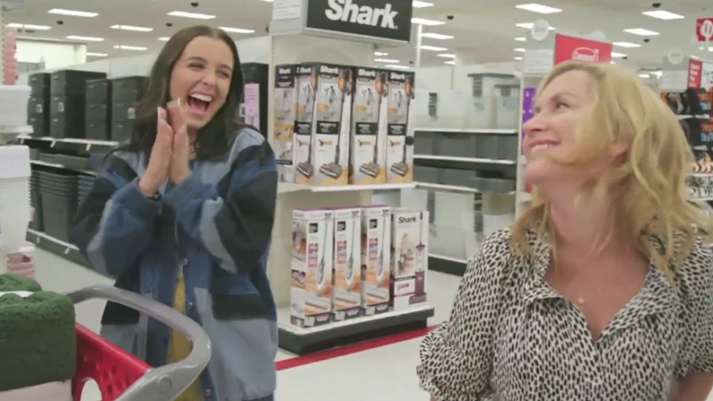Two adult females stand inside a retail store, one clapping and smiling with a shopping cart, the other looking towards the first.