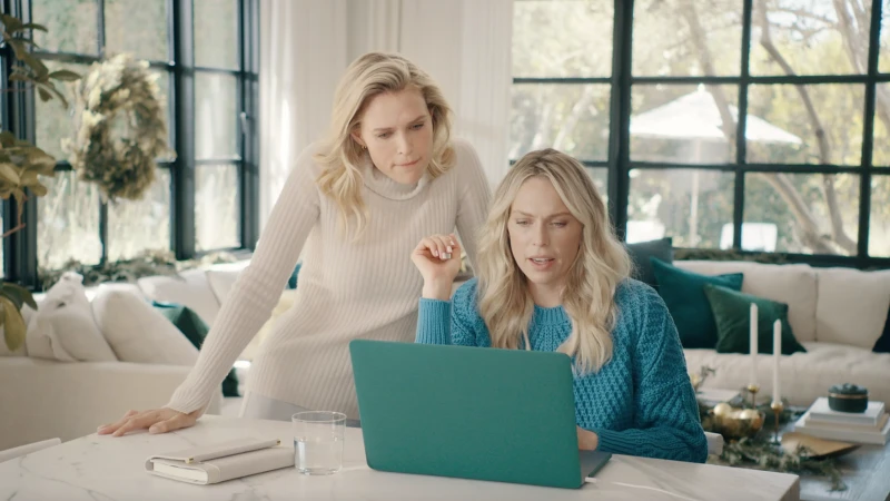 Two blonde women collaborate indoors at a table with a laptop, notebook, and glass; natural light filters through the large windows.