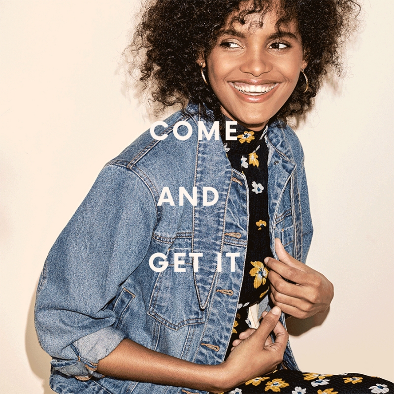 Smiling woman with curly dark hair wearing a denim jacket and floral top against a plain light background