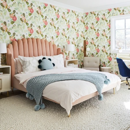 A furnished bedroom featuring a pink velvet scalloped headboard bed, floral wallpaper, and a pale blue knit throw blanket.