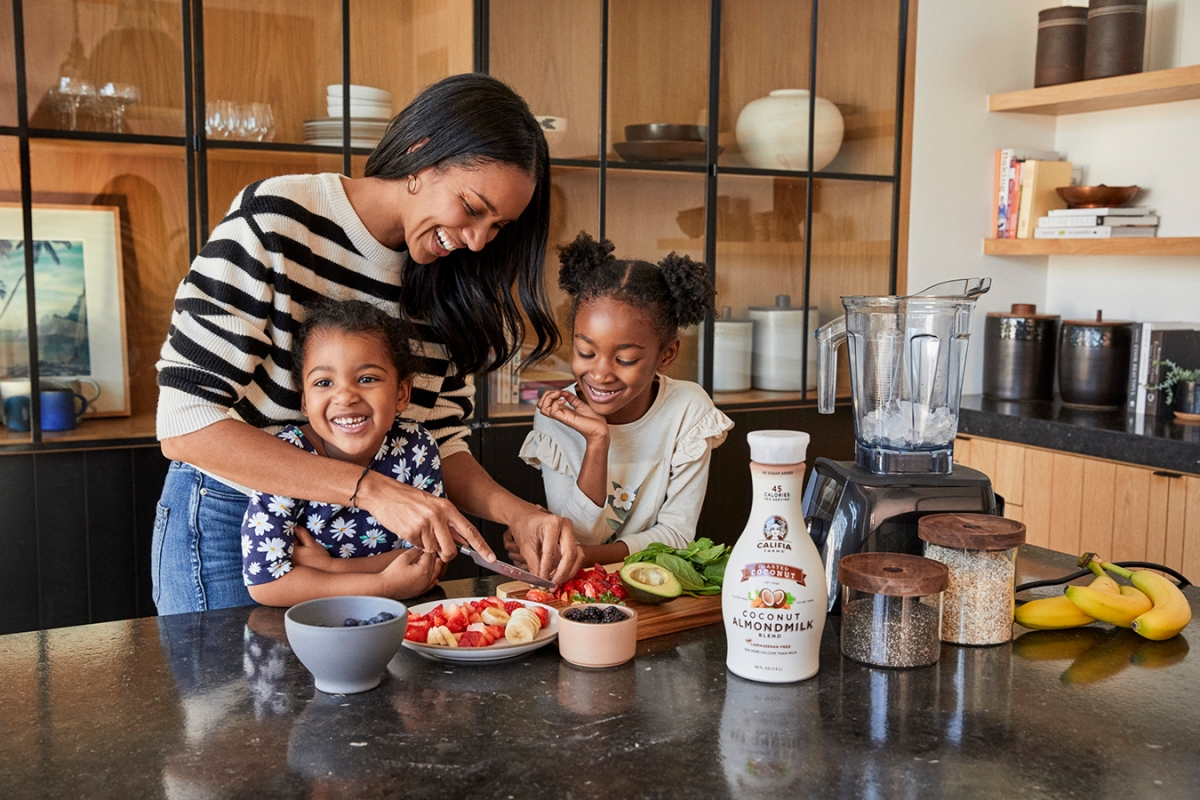 A smiling woman and two young girls are in a kitchen, cutting fruit for a smoothie, with a blender and ingredients on a countertop.